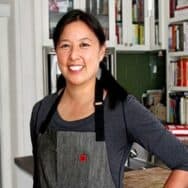 Michelle Tam standing with a gray apron in front of a bookcase, smiling