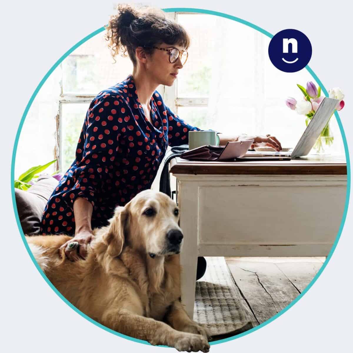 A woman sitting at a desk using a laptop, with a golden retriever lying by her side.