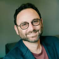Andrew Wilder, sitting on a couch wearing a casual blazer and t-shirt, smiling at you.
