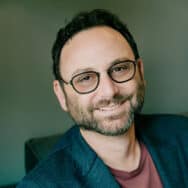 Andrew Wilder, sitting on a couch wearing a casual blazer and t-shirt, smiling at you.