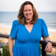A woman with brown hair in a blue dress standing in front of the ocean
