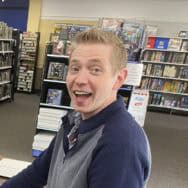 a caucasian man with blonde hair standing in a book store. he is wearing a blue and gray shirt and he is smiling.