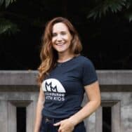 A smiling brunette woman wearing a gray shirt that says Milwaukee With Kids stands in front of a stone railing and trees.