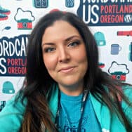 Amber Pittman, with long dark hair and a teal jacket, smiling in front of a WordCamp US backdrop.